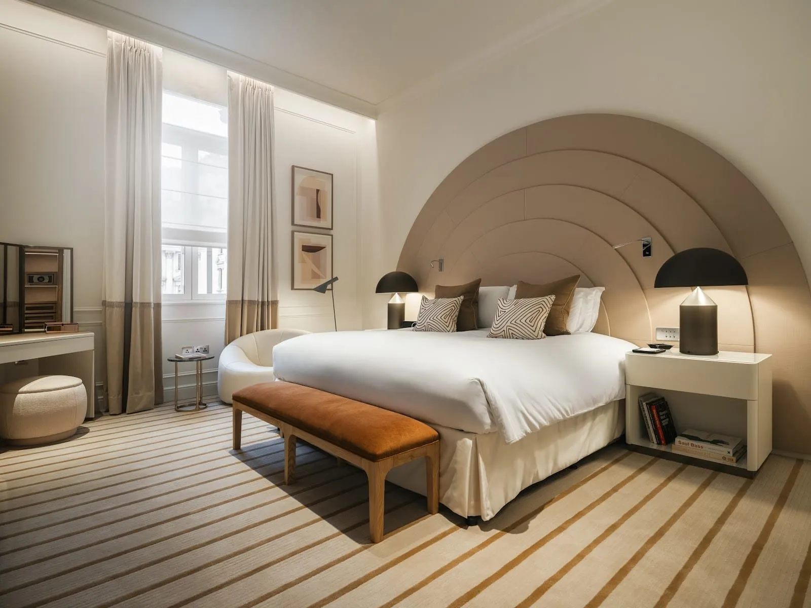 A room at Sofitel London St James with a huge semicircular headboard in stepped beige leather, black mushroom lamps, a striped carpet, a round white armchair and a tall window