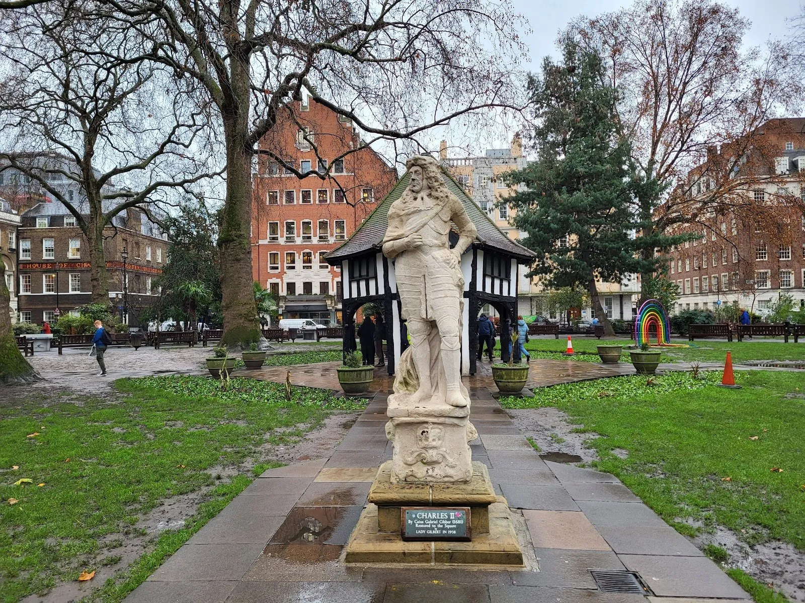 The weathered stone statue of Charles II on its plinth in Soho Square, a paved path leading past it to the half-timbered hut