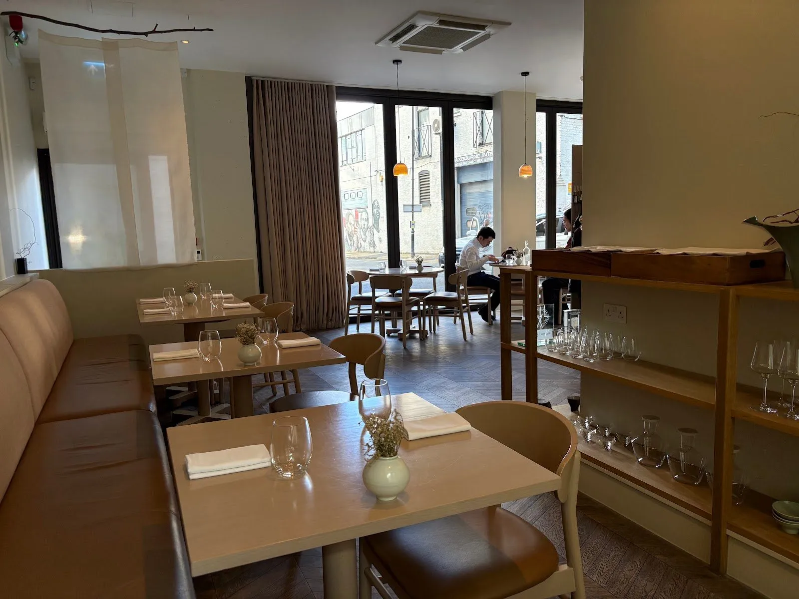 Sollip's pale dining room, with a long beige banquette, oak tables set with small vases of dried flowers, oak shelves of glassware and a glazed front onto Melior Street