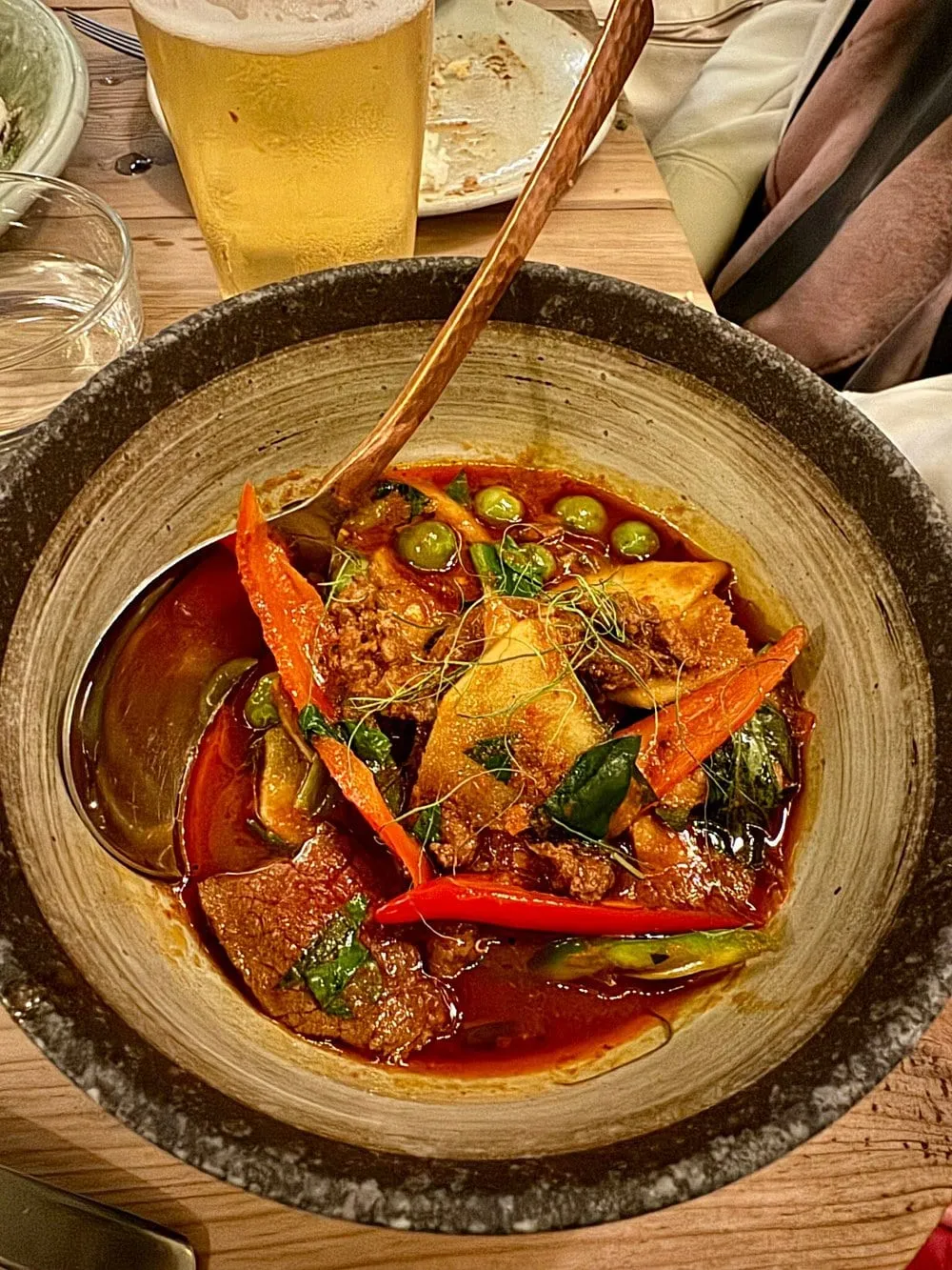 A red curry with beef, Thai aubergines, pea aubergines, whole red chillies and holy basil in a rough stoneware bowl, with a copper spoon and a glass of beer on the table