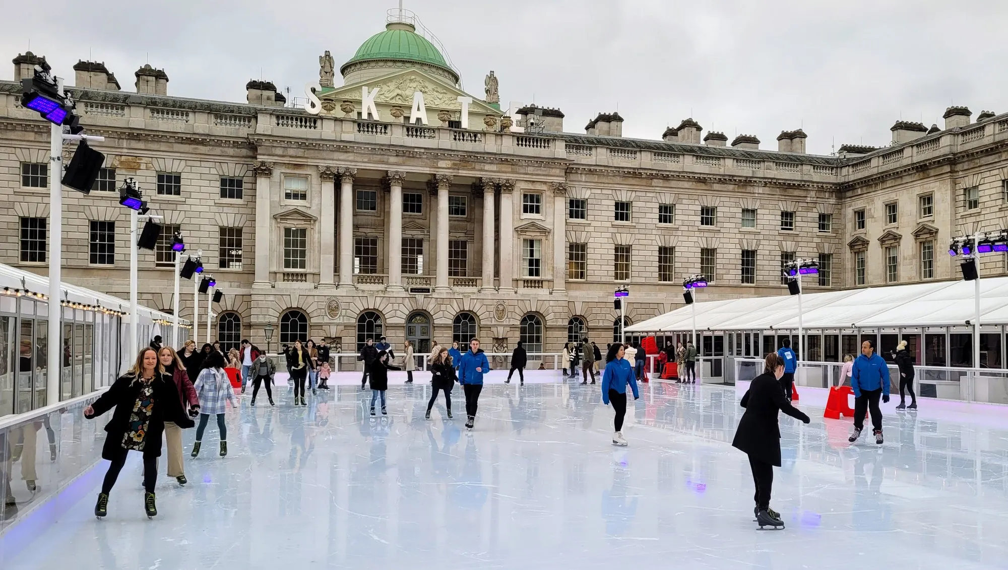 Skaters on the ice rink in the courtyard of Somerset House, with SKATE spelled in illuminated letters along the roofline of the neoclassical building behind