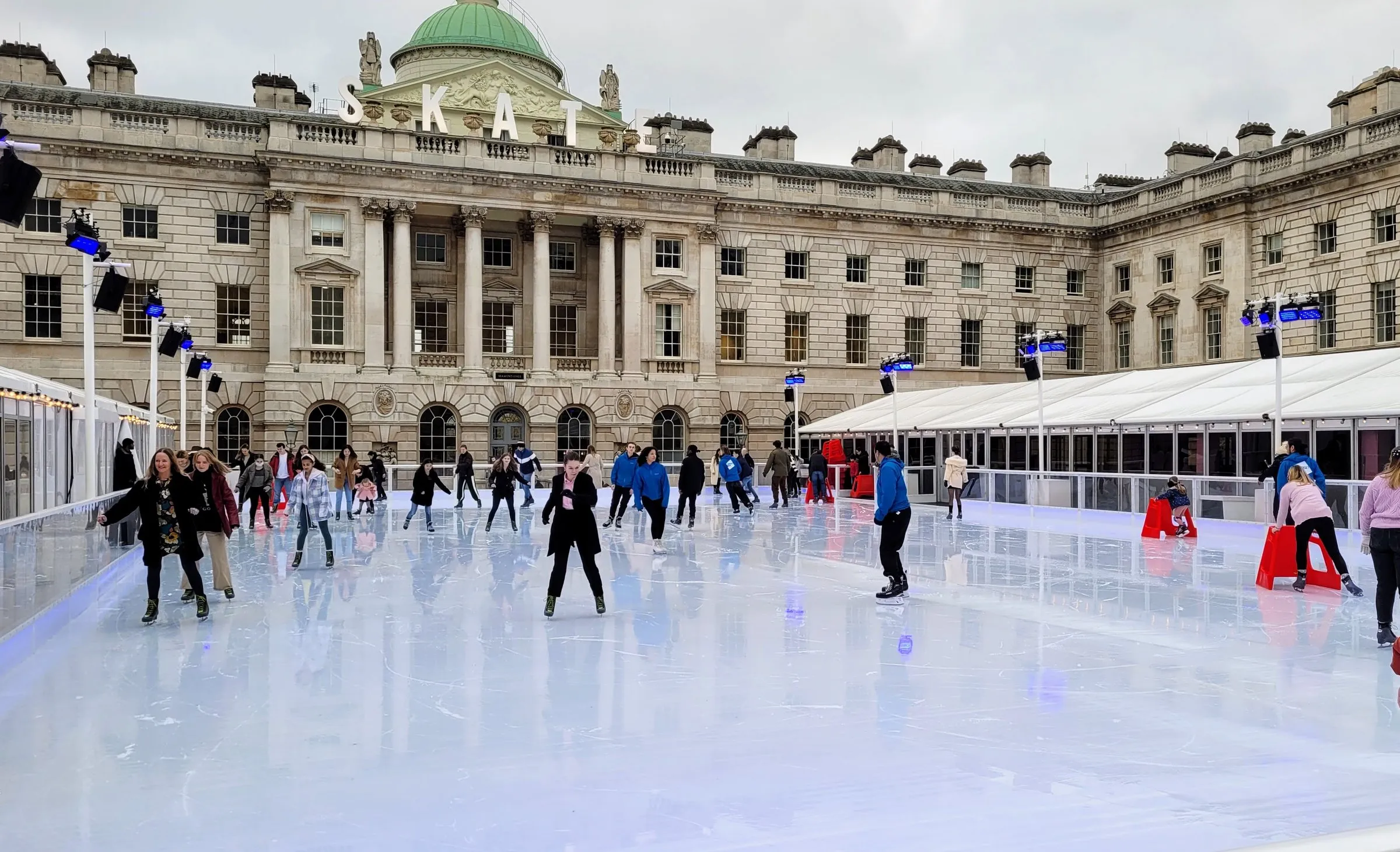 Skaters on the Somerset House ice rink on an overcast day, in front of the domed courtyard front with large white SKATE letters along its roofline and a marquee down the right