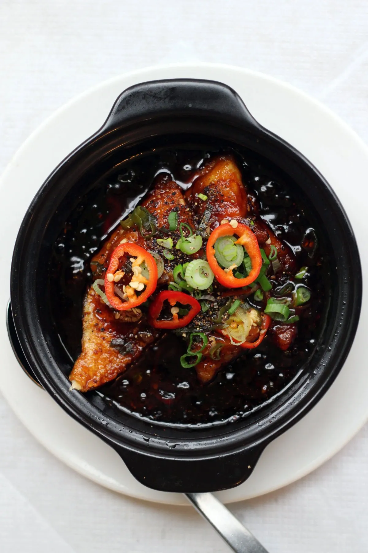 A clay pot of caramel-braised fish topped with sliced red chilli and spring onion, photographed from above on a white plate