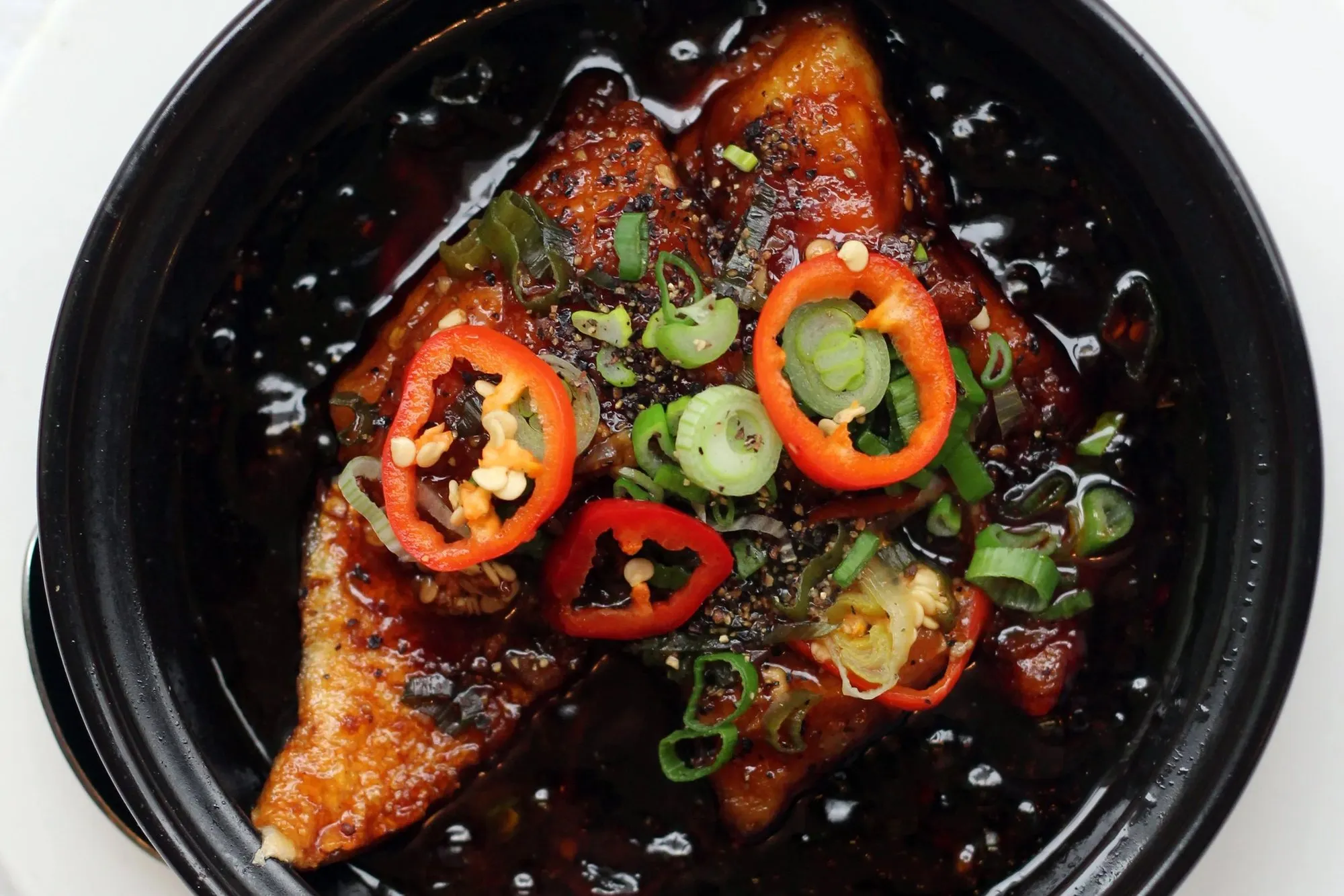 A clay pot of caramel-braised fish topped with sliced red chilli and spring onion, photographed from above on a white plate