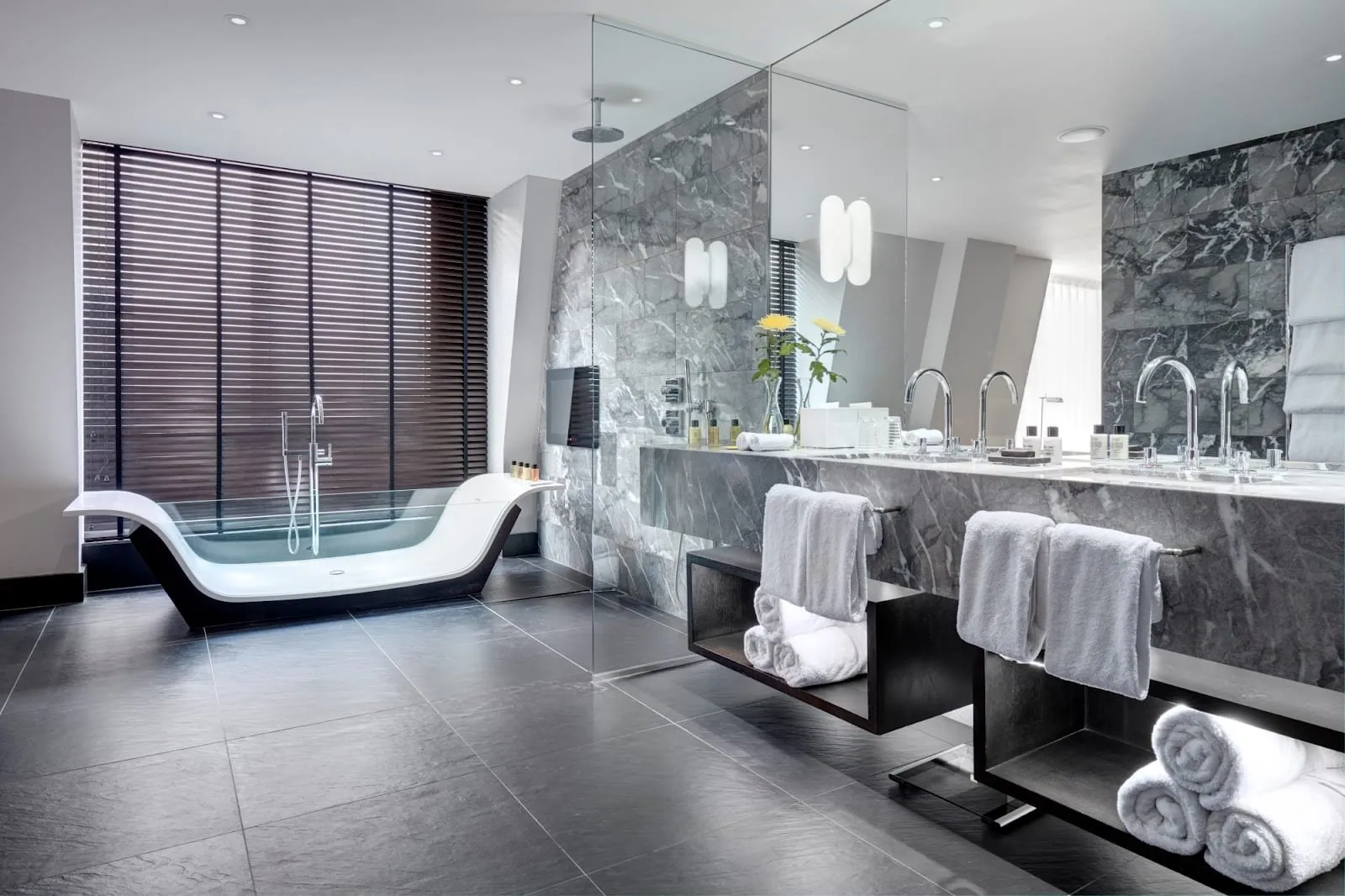 A suite bathroom at South Place Hotel with a glass-sided freestanding bath under slatted blinds, a walk-in rain shower and two basins in grey marble
