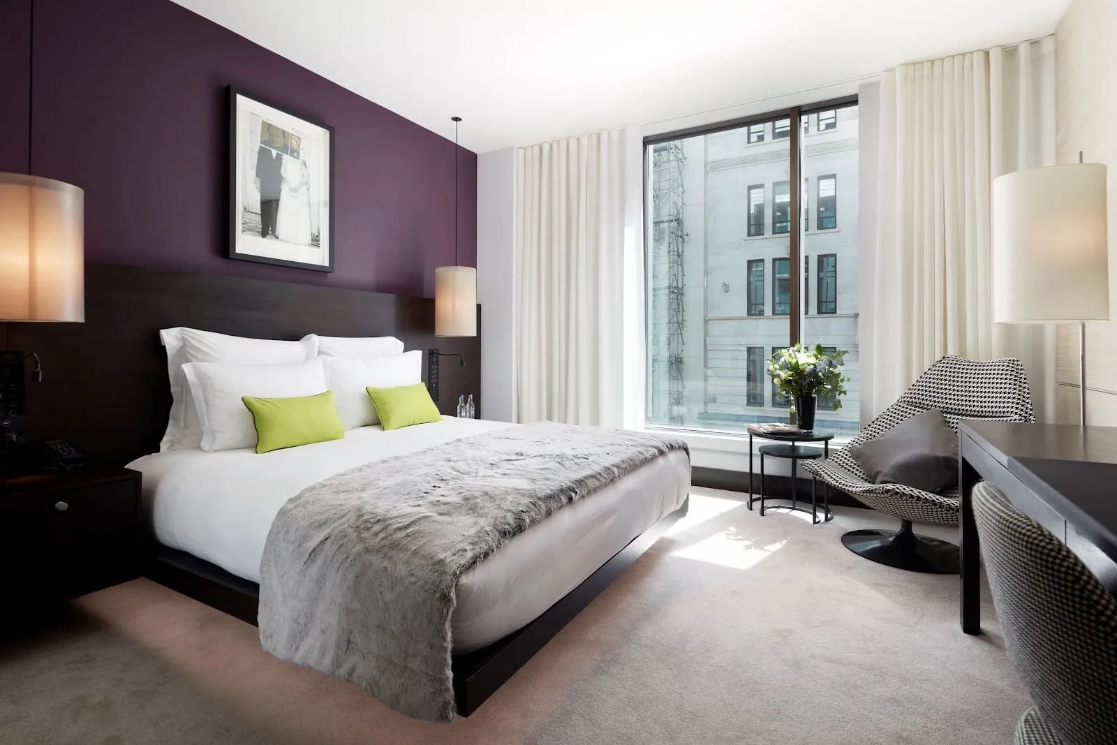 A double room at South Place Hotel with an aubergine feature wall, a dark wood headboard, lime cushions, a grey fur throw and a floor-to-ceiling window onto the stone building opposite