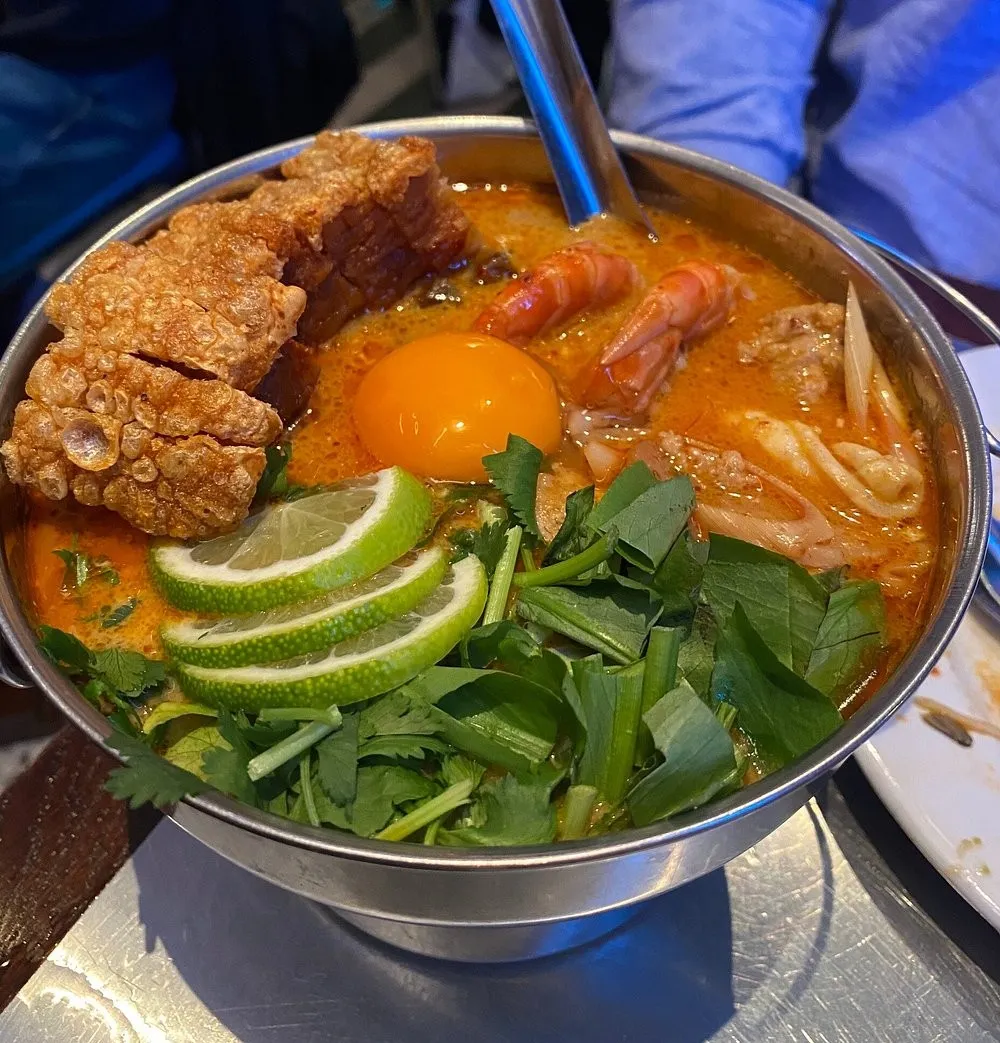 A metal pot of orange-red soup with prawns, crisp pork belly, a raw egg yolk, lime slices and coriander
