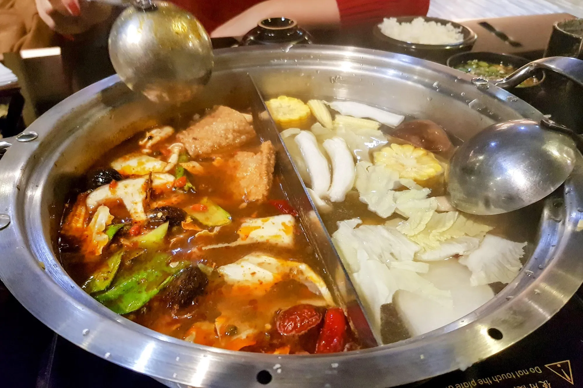 A divided steel hot pot on a table burner, one half red with chilli and Sichuan pepper, the other a clear broth with cabbage, corn and rice cakes