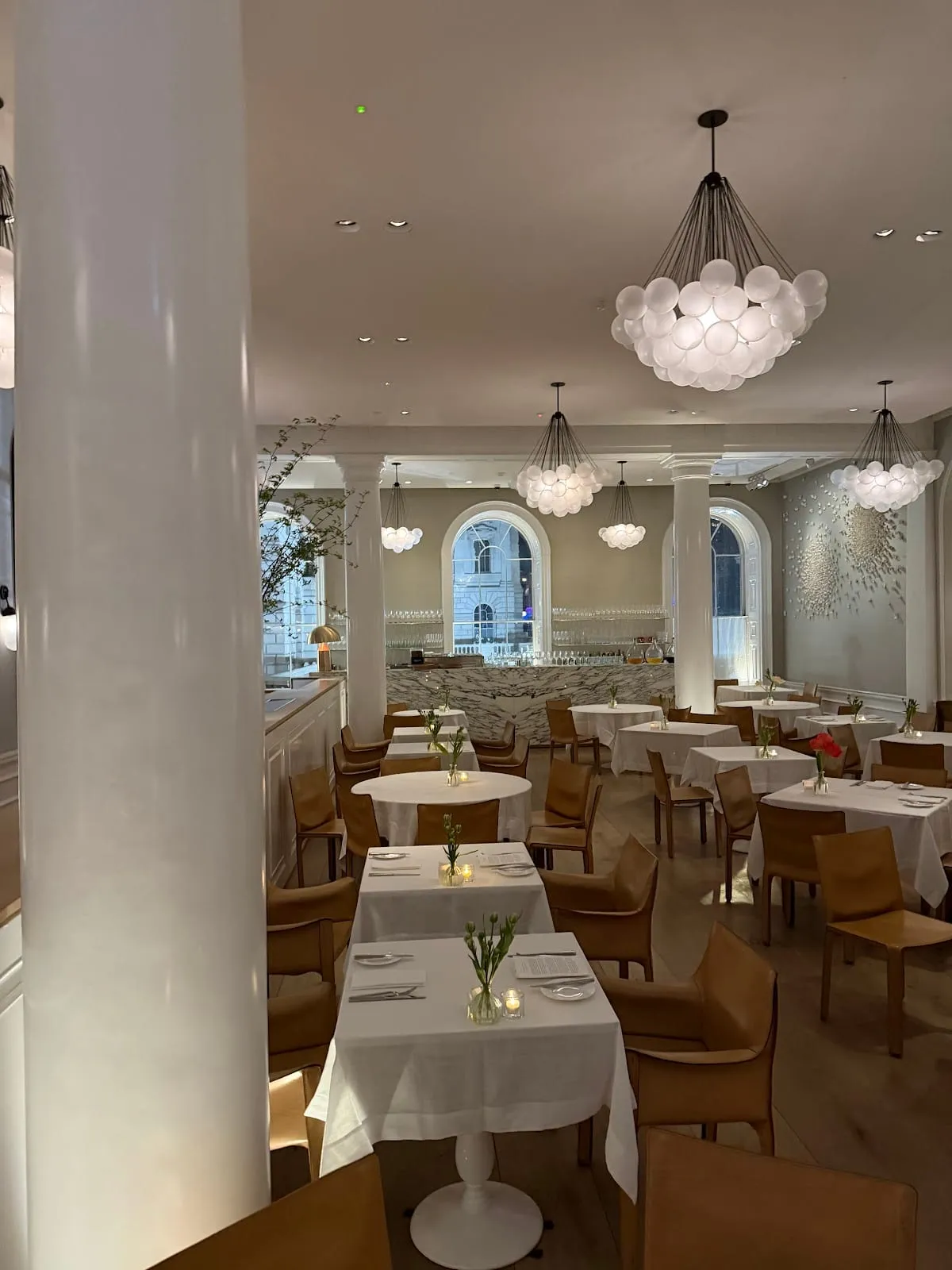 The dining room at Spring in Somerset House, white-clothed tables and tan leather chairs between white columns, under clusters of white globe lights