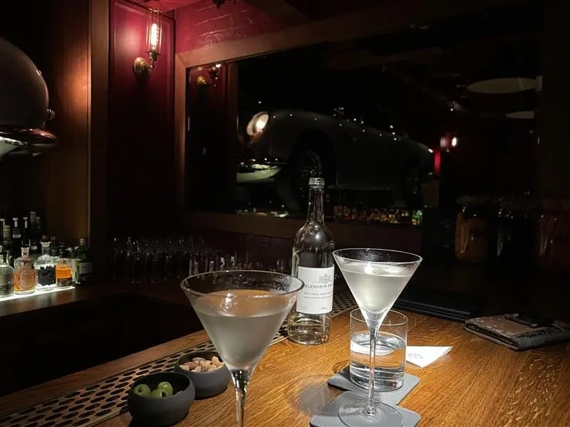 Two martinis on a wooden bar counter at The Spy Bar, with olives, a water bottle and dark red walls behind