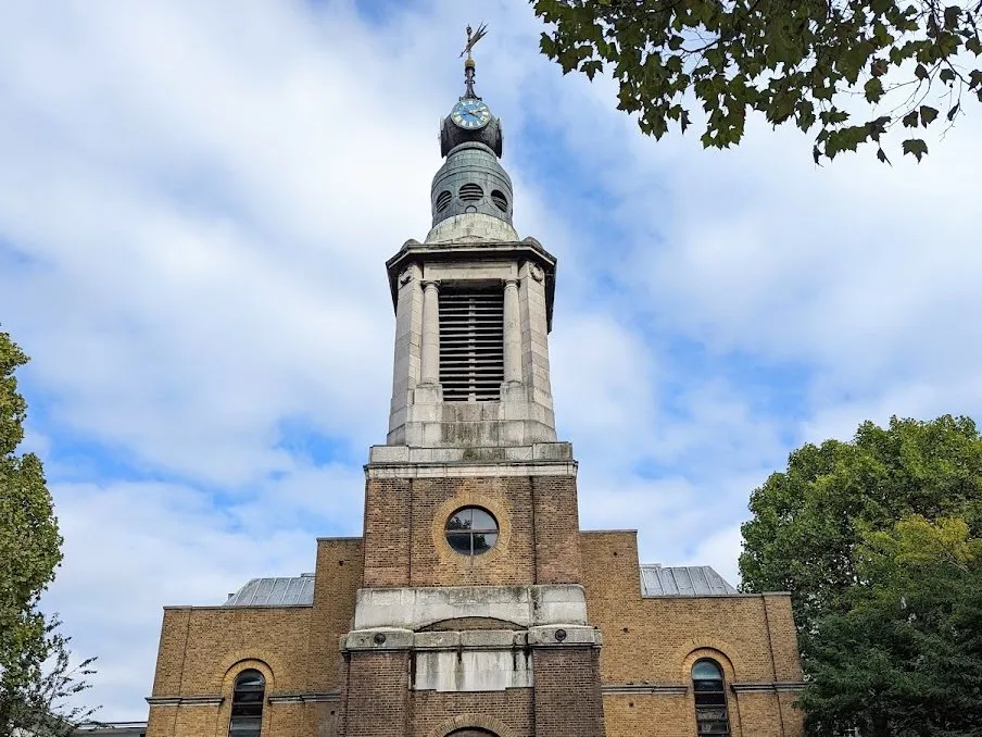The tower of St Anne's Soho: a brick base with a round window rising to a stone belfry, a copper-clad drum and a blue clock face, with a weathervane on top