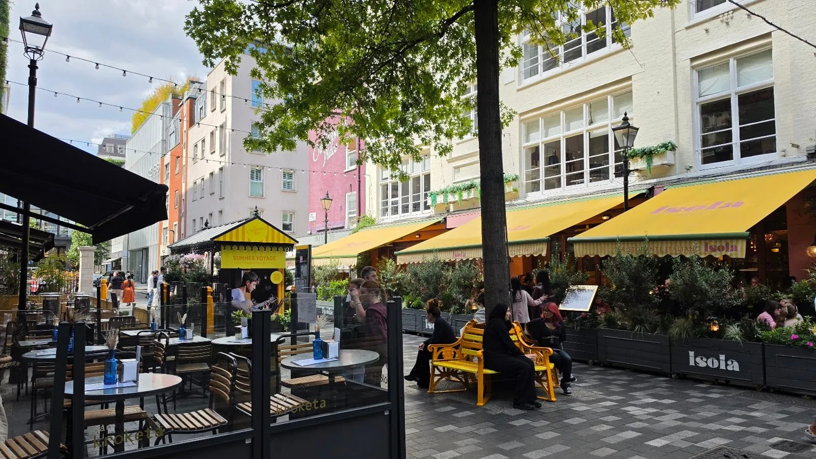 Outdoor café tables under yellow awnings along the pedestrianised St Christopher's Place, with shops and string lights above