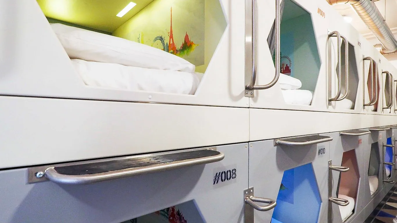 A wall of white moulded sleeping capsules stacked two high, numbered 007 to 010, each with a hexagonal window, a steel grab handle and a painted city mural inside