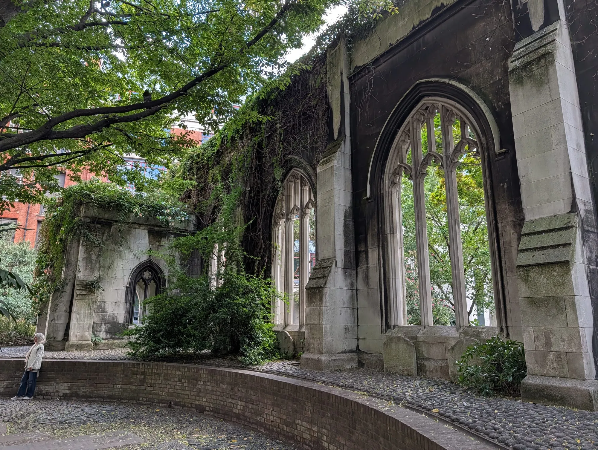 The ruined shell of St Dunstan-in-the-East, Gothic window tracery standing open to the sky with ivy grown over the walls and a tree leaning across the cobbled path