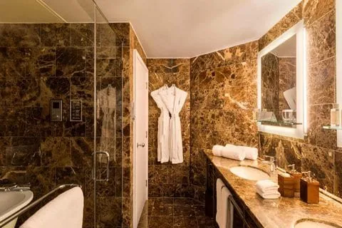 A bathroom at St Ermin's Hotel lined in dark brown marble, with a glass-walled shower, twin basins under a lit mirror and a white robe on the door