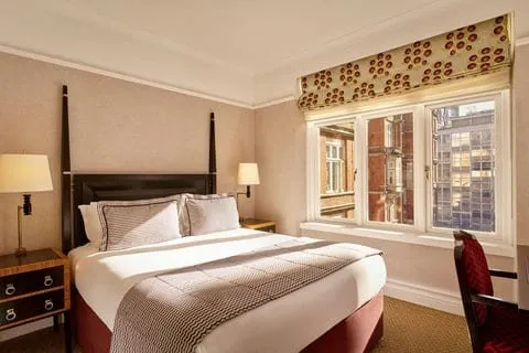 A double room at St Ermin's Hotel with a dark wooden four-poster-style bed frame, a houndstooth throw, and a wide window with a patterned blind looking onto red-brick buildings