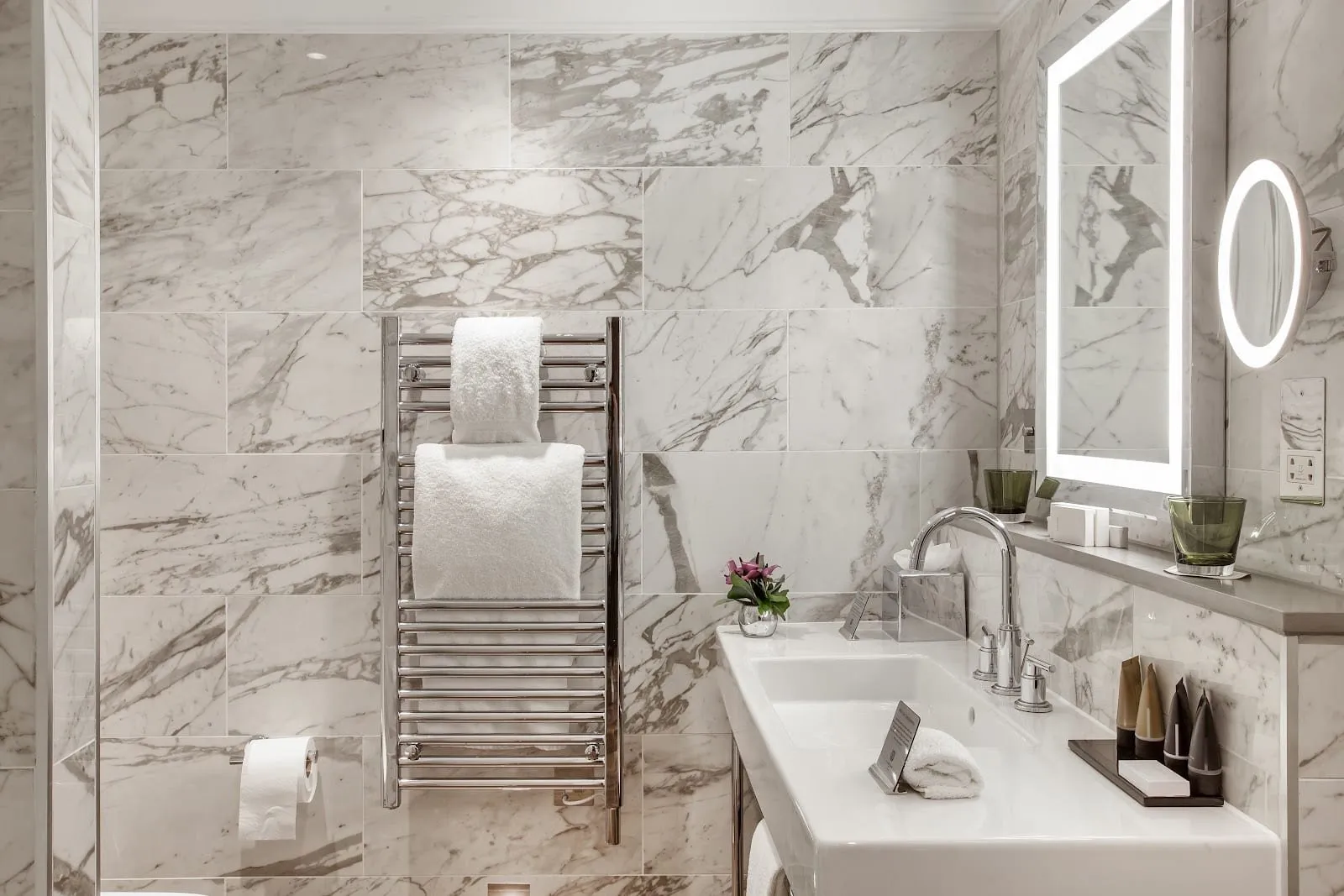 A bathroom at St. James' Court in white and grey veined marble, with a heated towel rail, a white basin on a chrome stand and a backlit mirror with a round make-up mirror