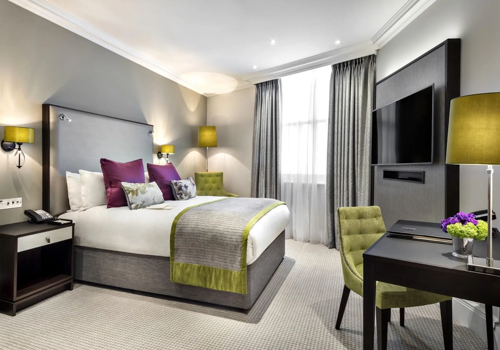 A double room at St. James' Court with a tall grey headboard, purple and floral cushions, lime-green velvet chairs, lime lamp shades and a TV set into a fabric panel