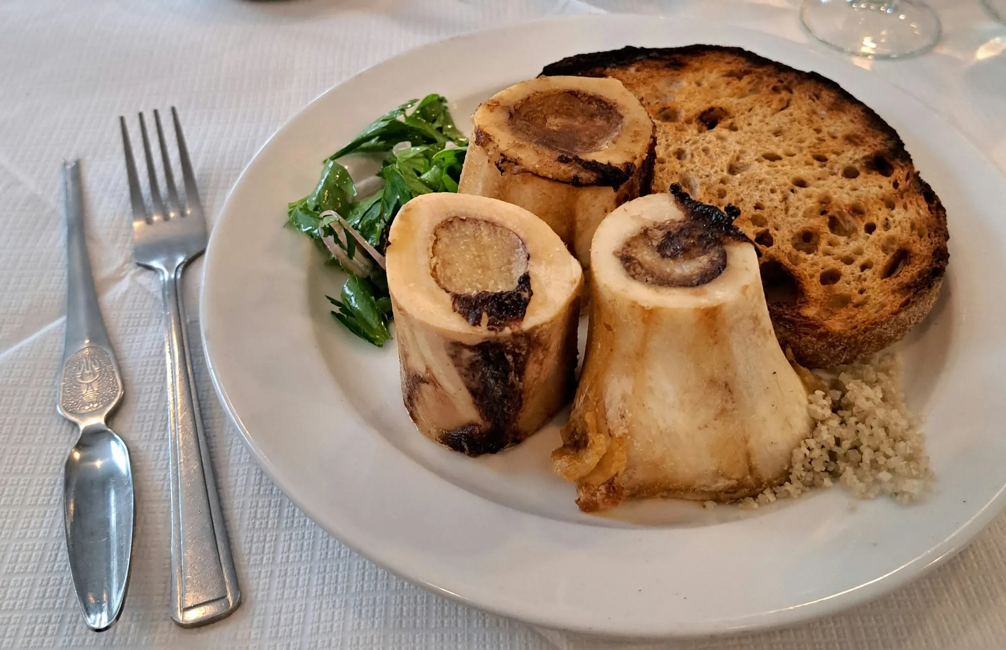 Three roasted bone marrow halves on a plate with a wedge of toast and a parsley salad.