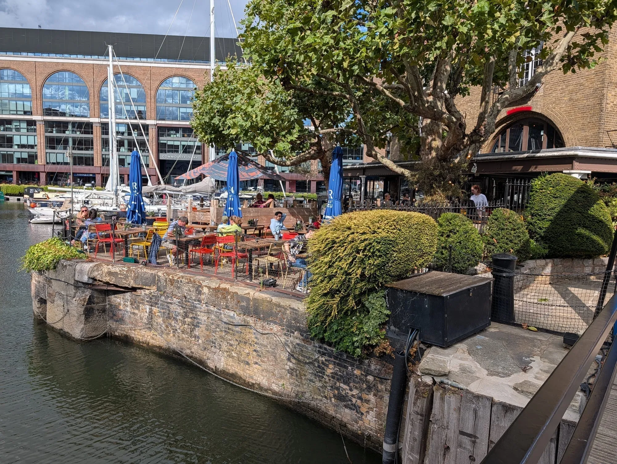 Tables and bright red and yellow chairs on a stone dock wall at St Katharine Docks, moored yachts alongside and the brick arches of Commodity Quay behind