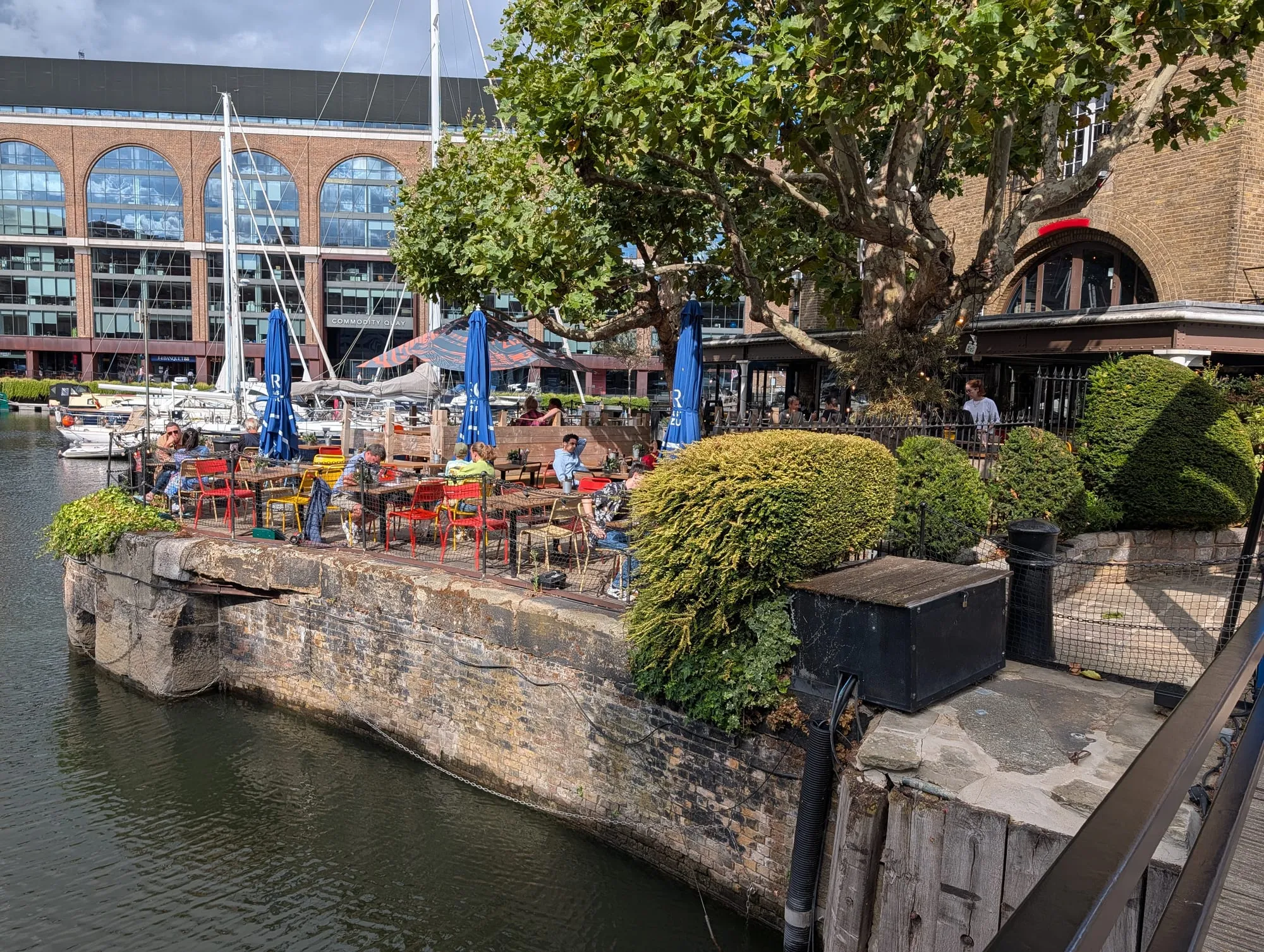 Tables and bright red and yellow chairs on a stone dock wall at St Katharine Docks, moored yachts alongside and the brick arches of Commodity Quay behind