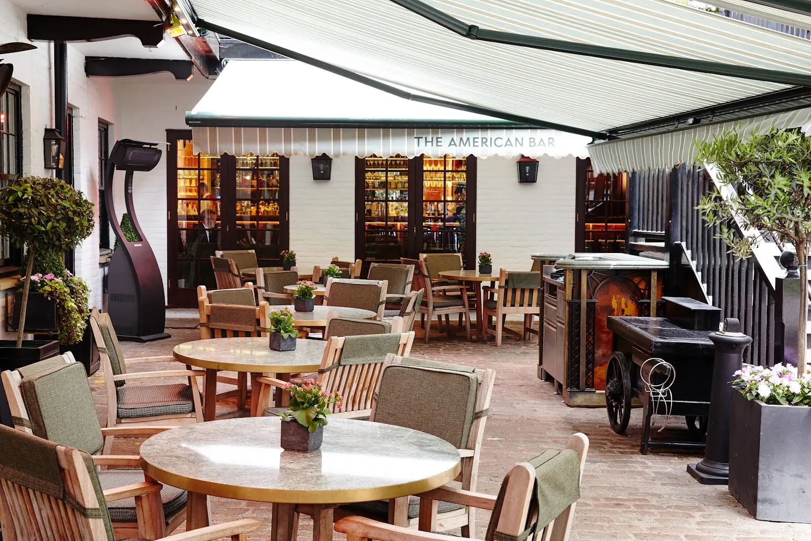 The cobbled courtyard at The Stafford, with wooden tables and cushioned chairs under striped awnings outside the American Bar, olive trees in planters and a heater between the tables