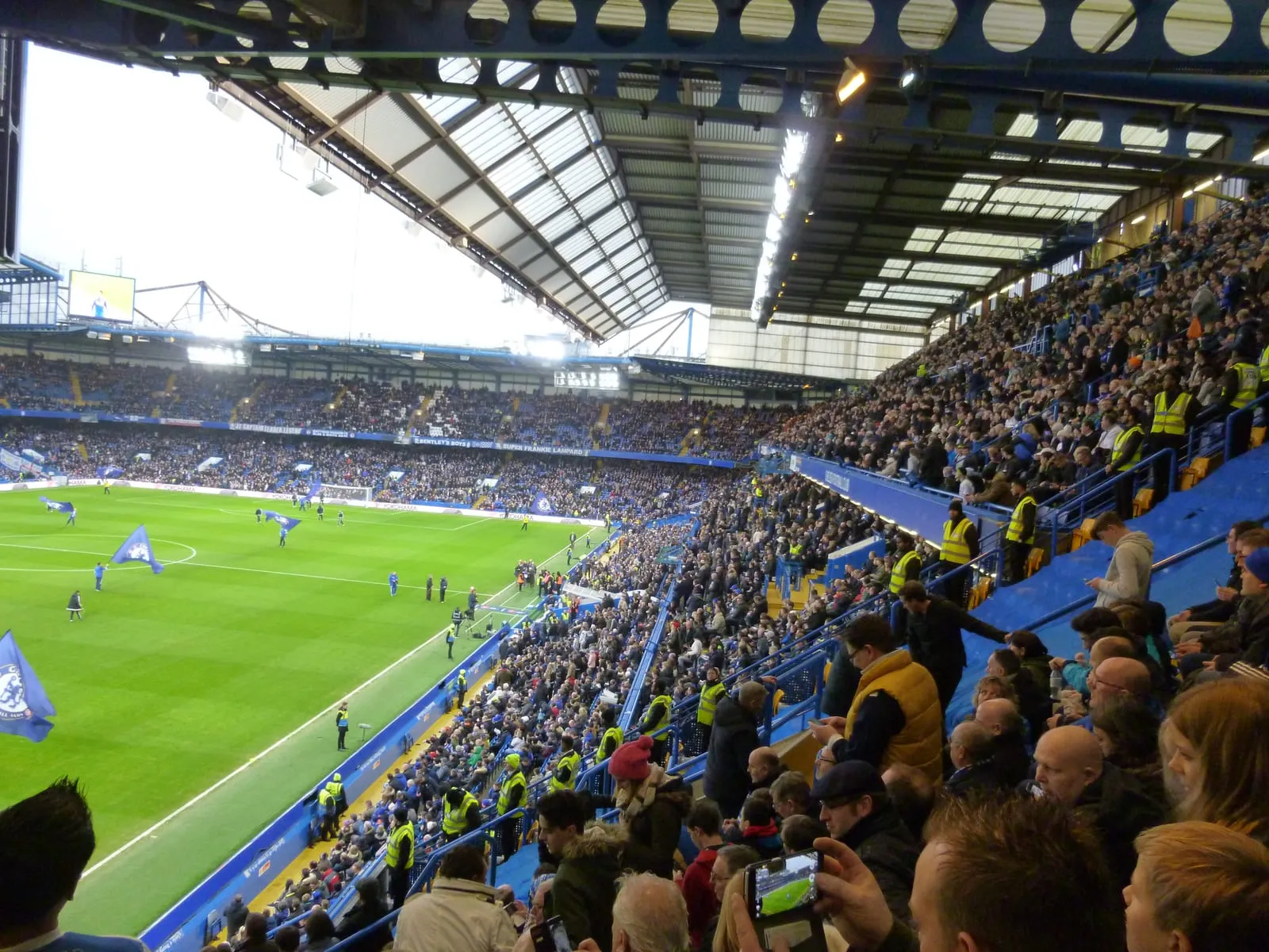 A packed Stamford Bridge seen from high in the stand, supporters banked steeply above the pitch with stewards in hi-vis lining the front rows