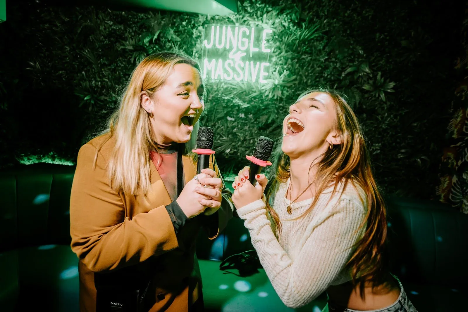 Two women singing into microphones in front of a leafy wall with a neon sign reading Jungle Massive