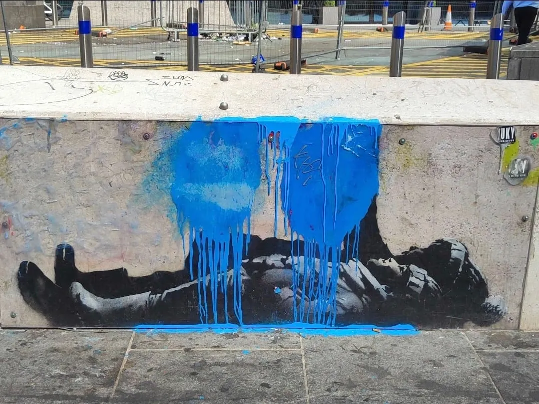 Banksy's stencil of two children lying on their backs on a low stone wall at Centre Point, splashed with blue paint that has run down over both figures