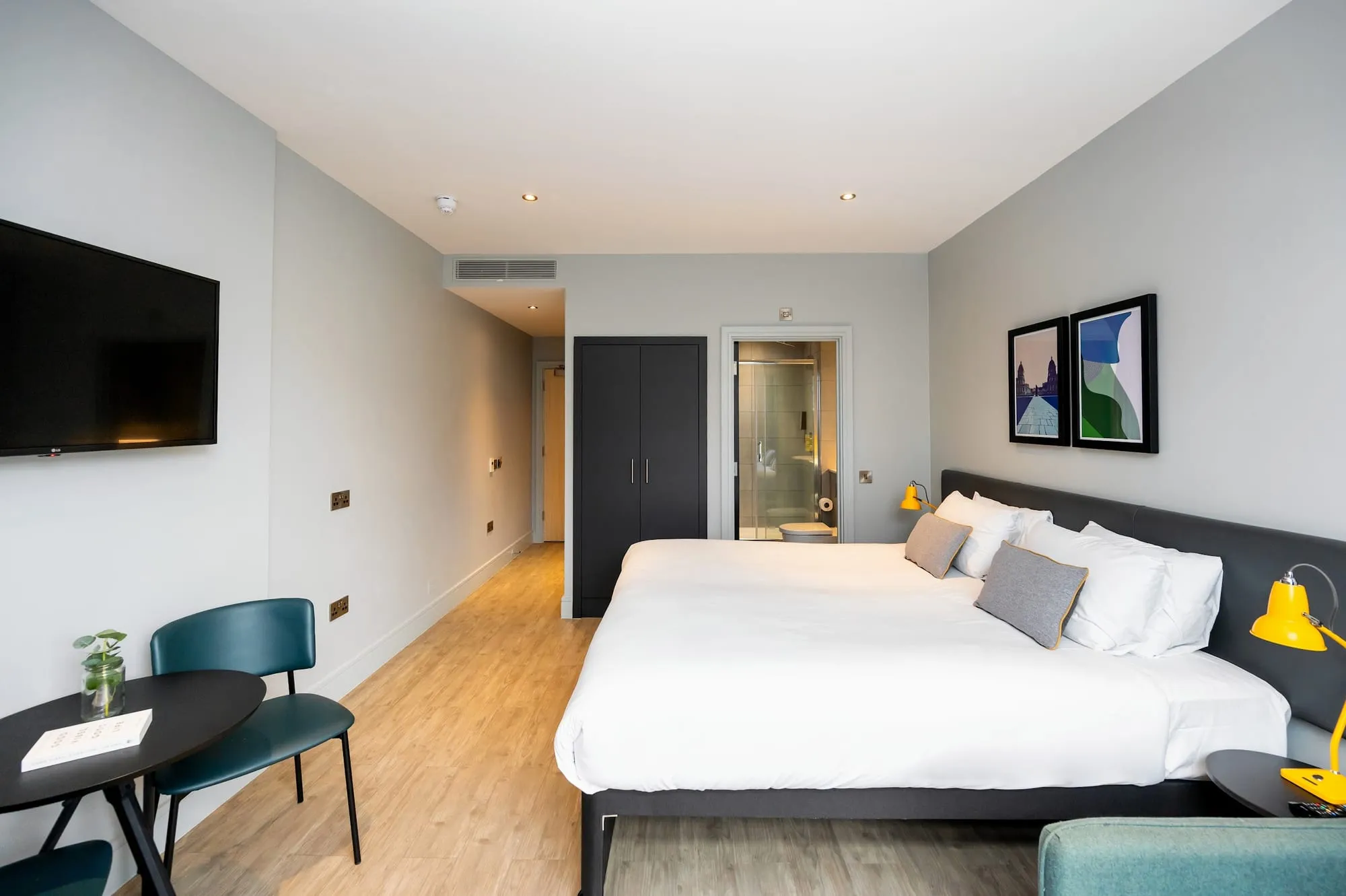 A bedroom at Staycity Greenwich High Road, with a wall-mounted television, a dark wardrobe and the ensuite bathroom visible through an open door