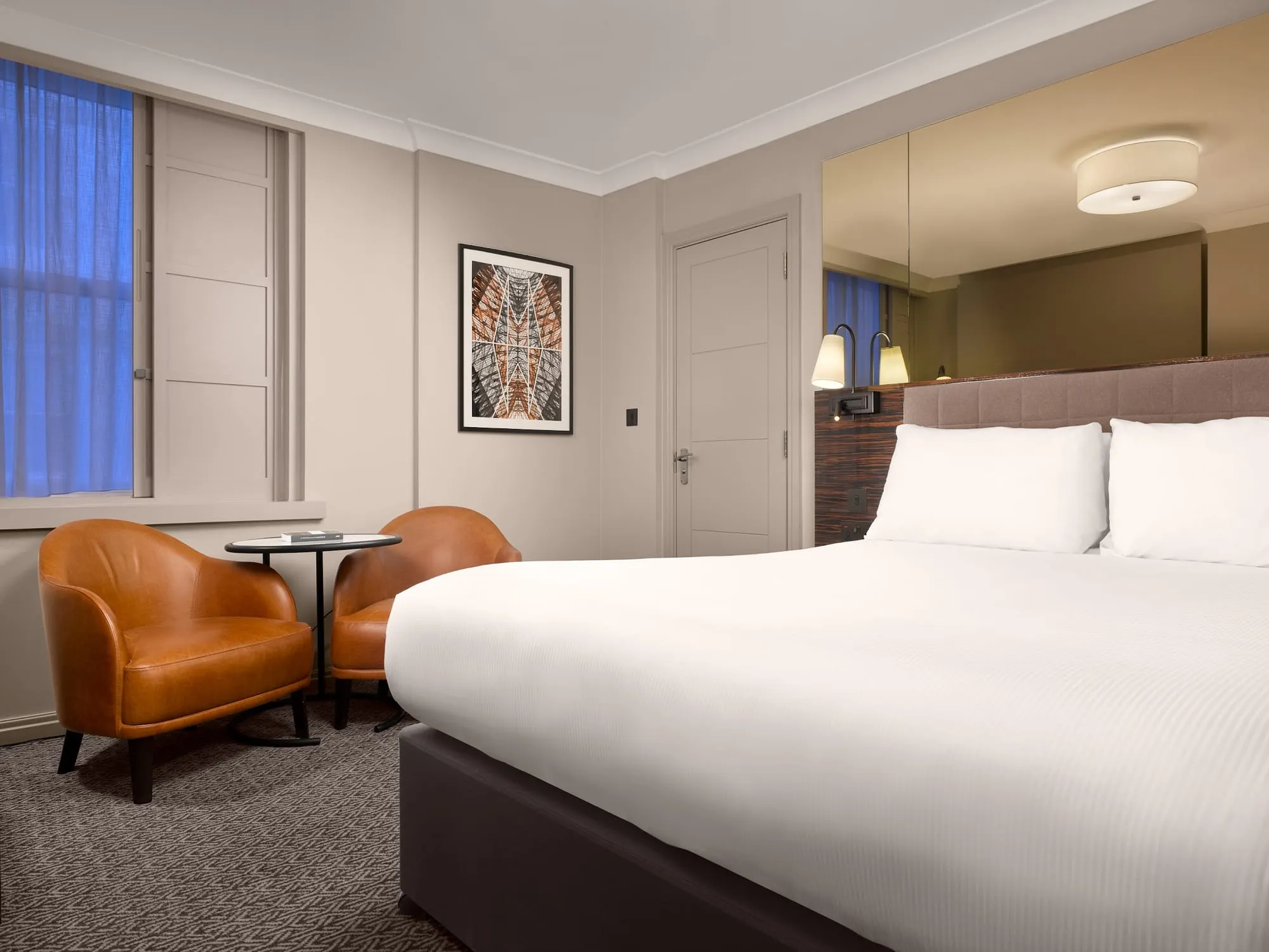 A Strand Palace guest room with white bedding, two tan leather armchairs around a round table, and a mirrored panel behind the headboard