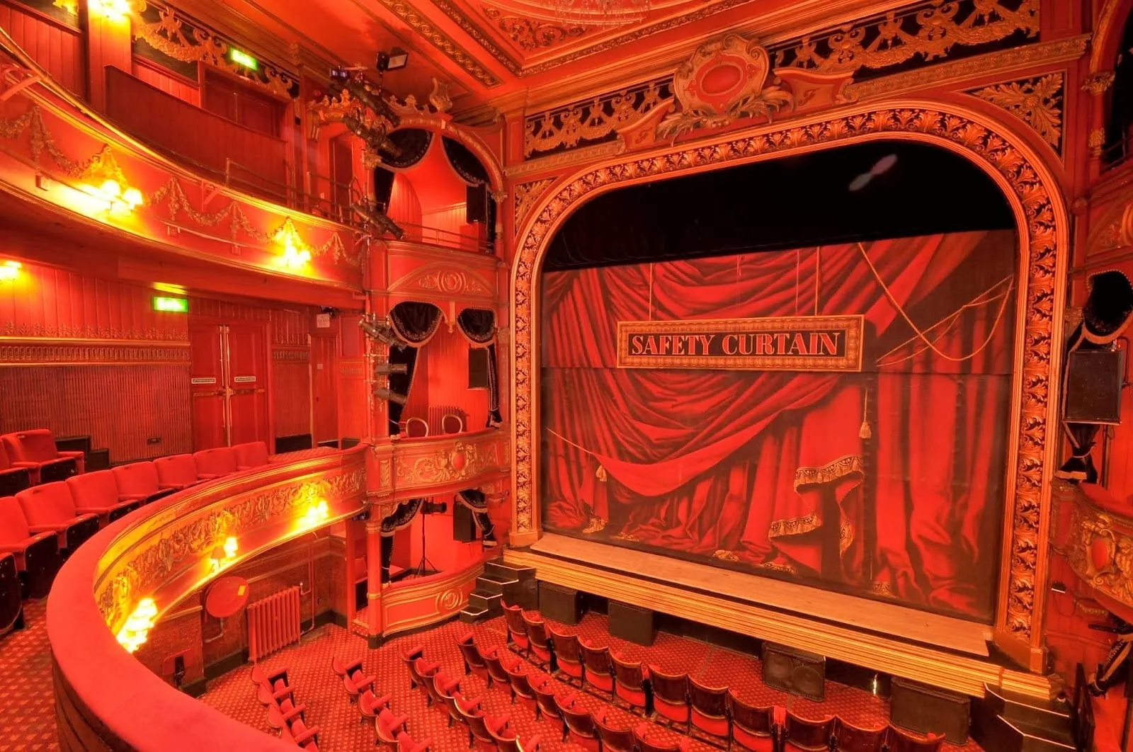 The red and gold auditorium of Theatre Royal Stratford East, with a curved circle, stage boxes and a safety curtain painted as red drapes