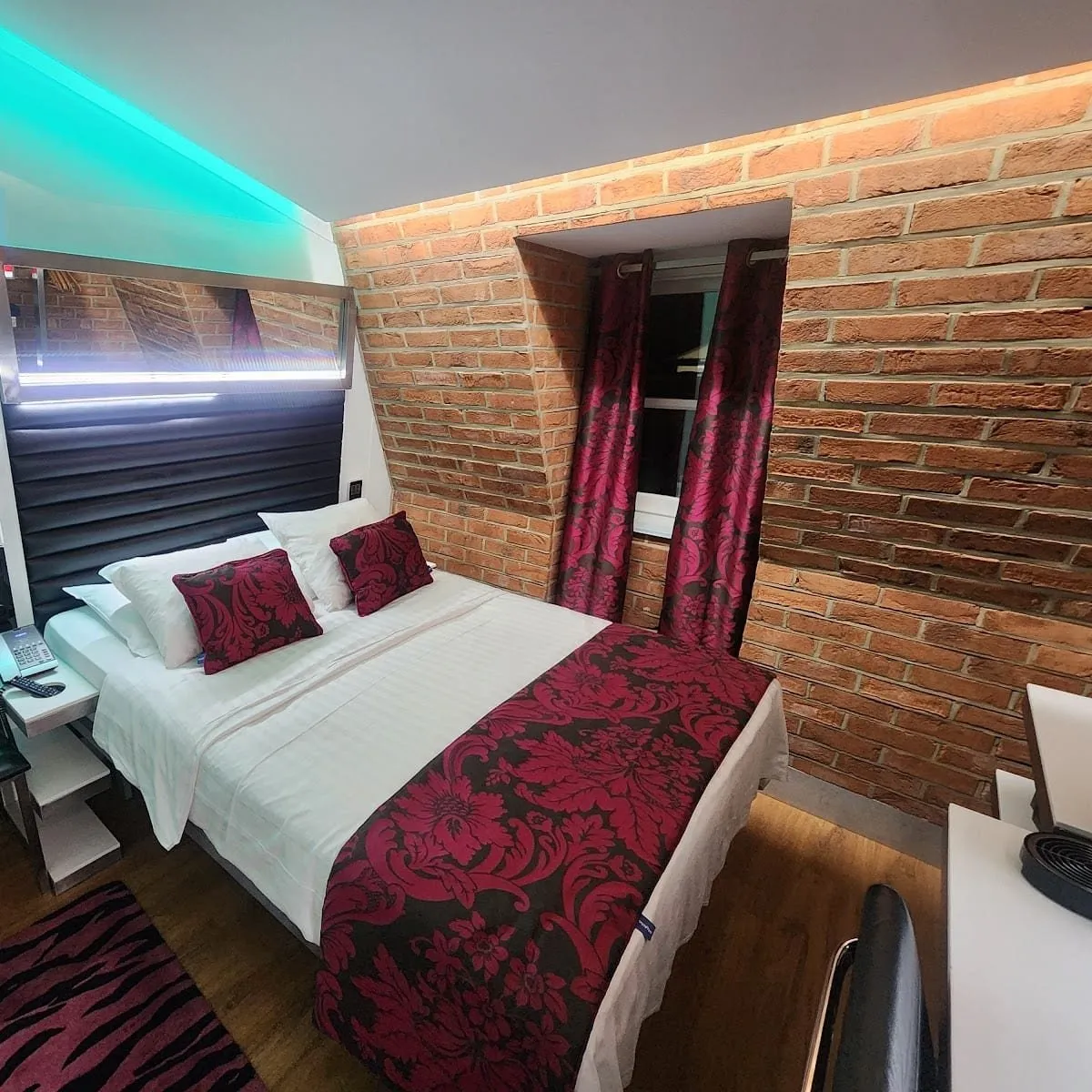 A double room at Stylotel under a sloping ceiling, with exposed brick walls, a small window with red damask curtains, a black padded headboard and a strip of turquoise light overhead