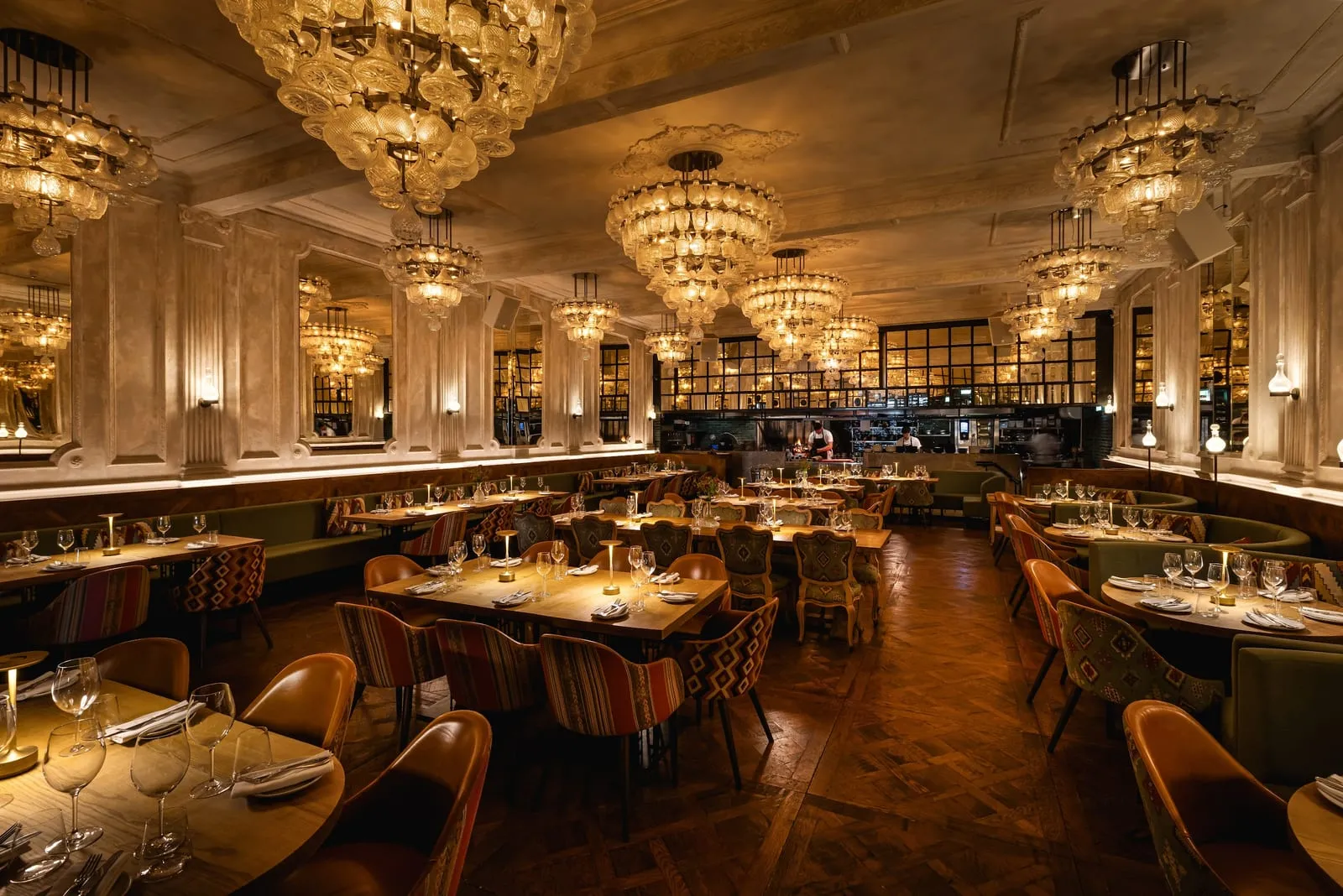 The long dining room at Sucre, clusters of glass chandeliers over leather chairs and banquettes, with the open kitchen lit at the far end