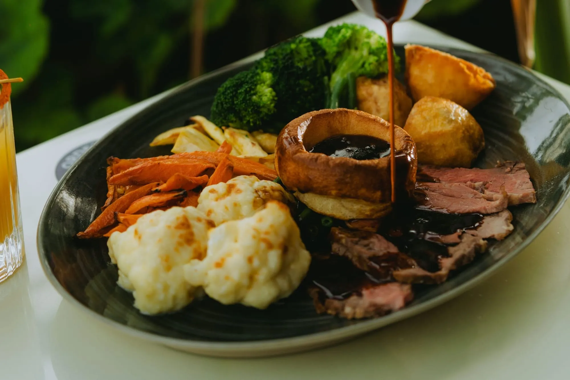 Gravy being poured into a Yorkshire pudding on a Sunday roast plate with beef, roast potatoes, cauliflower cheese and vegetables