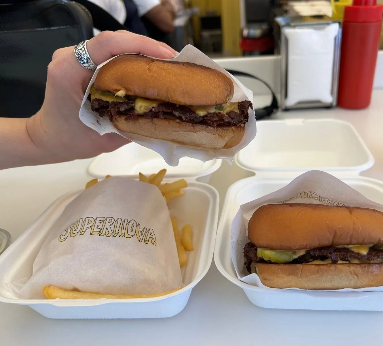 Two Supernova cheeseburgers in paper wrappers, one held up and one in an open takeaway box, each with a thin crisp-edged patty, pickles, melted cheese and house sauce, beside fries in paper printed with the Supernova name