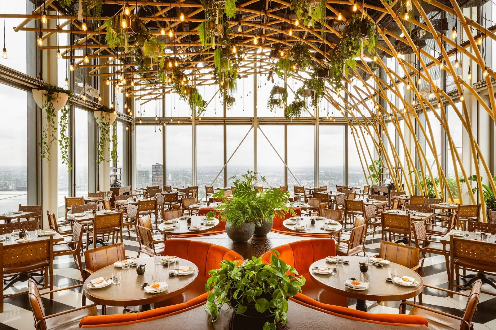 A high dining room on the 38th floor of Heron Tower, with arched bamboo poles and hanging plants overhead, orange velvet banquettes and floor-to-ceiling windows over London
