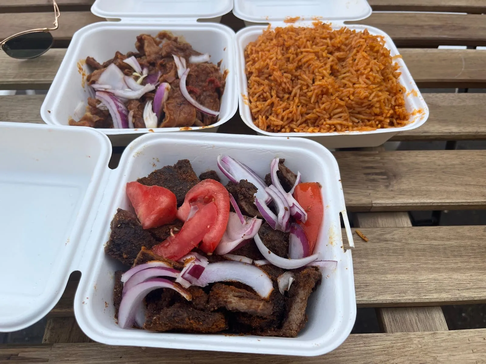 Three white takeaway boxes open on a wooden bench: thick slabs of dark, spice-crusted grilled beef under tomato wedges and raw red onion, thin-sliced beef in a dark sauce with more onion, and a box of orange jollof rice