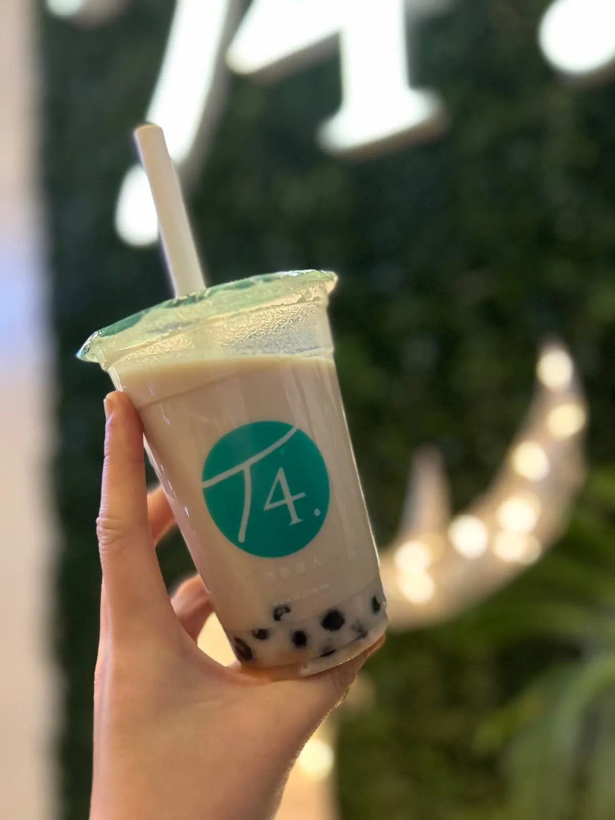 A T4 milk tea with dark pearls at the bottom, held up in front of a green wall with the T4 logo behind
