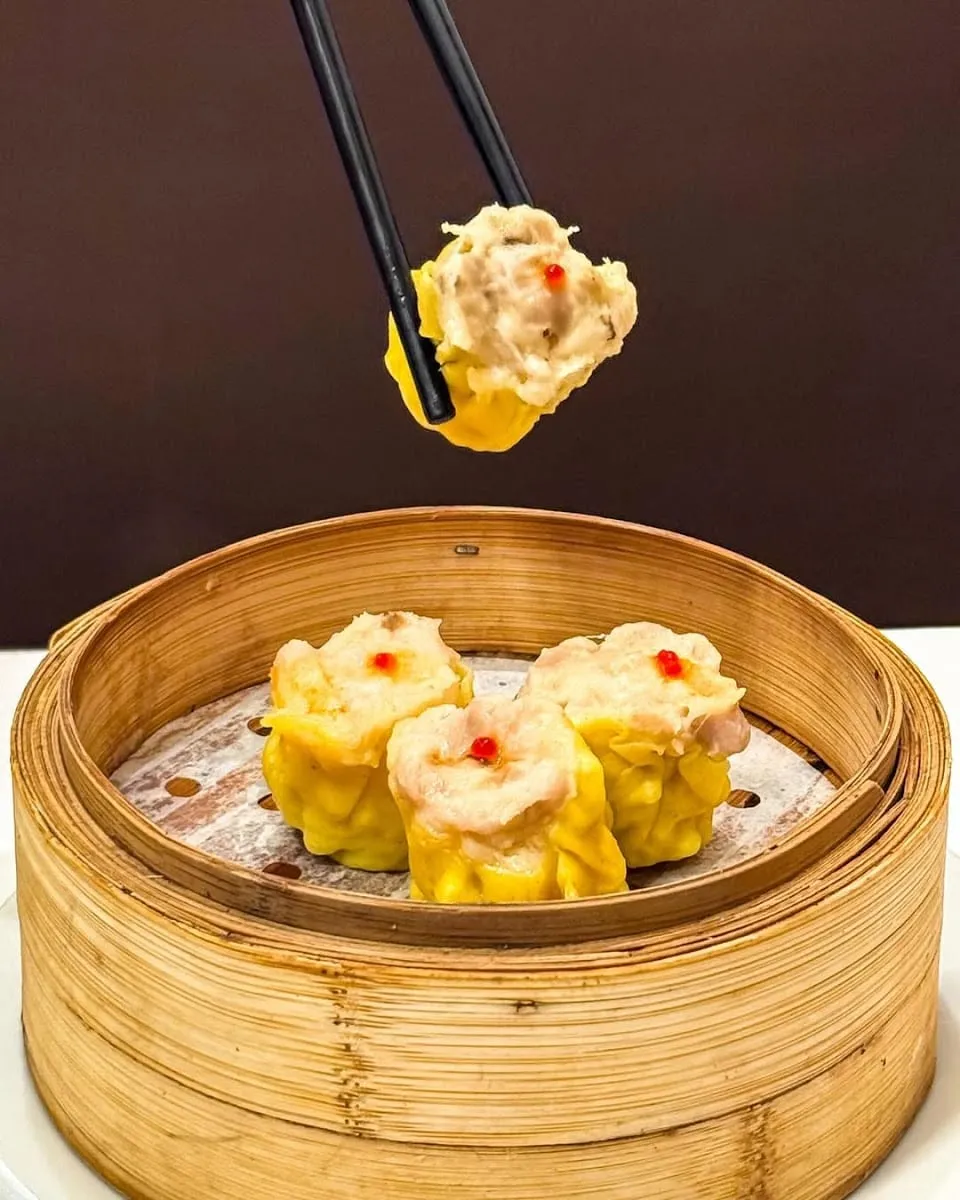 Three siu mai dumplings with pink pork and shrimp filling topped with a red dot, one lifted by chopsticks from a bamboo steamer