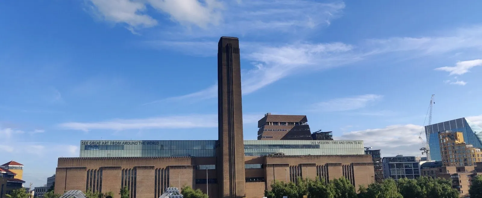 Tate Modern's brick power station and central chimney from the Millennium Bridge, with the stepped brick Blavatnik Building behind and a glass roofline reading See great art from around the world