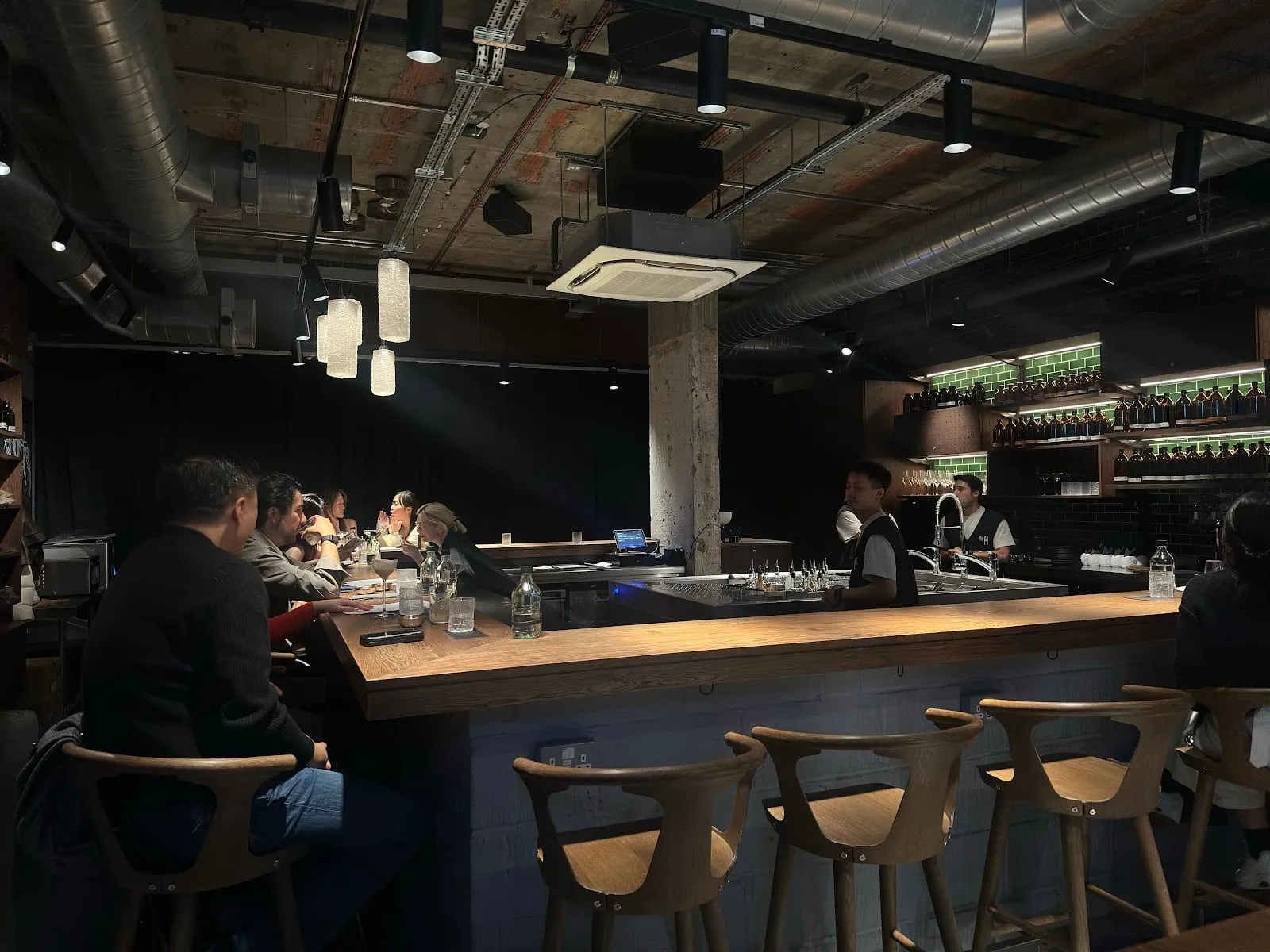 Customers on wooden stools at a long wooden bar counter, with bartenders working in front of shelves of amber bottles set against green tiles, under an exposed concrete ceiling