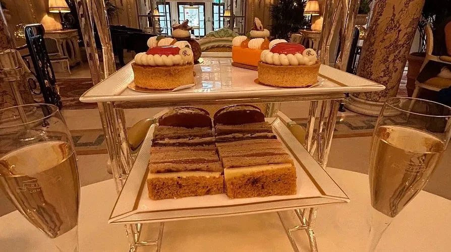 A tiered stand at the Ritz with finger sandwiches below and monogrammed pastries above, between two glasses of champagne