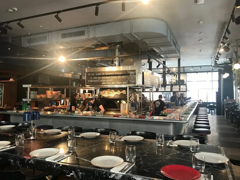 The open kitchen at temper seen across a marble counter laid for service, with chefs working by a wood-fired oven and a chalkboard above the pass