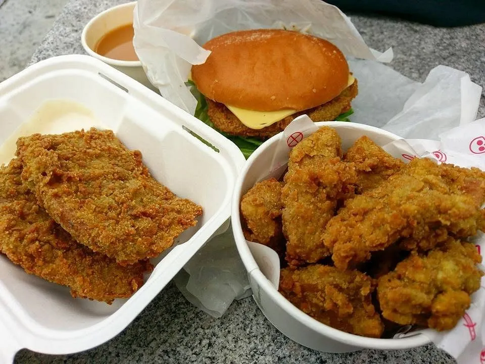 Seitan tenders in a takeaway box with a dip, a burger with melted cheese and lettuce, and a tub of fried popcorn pieces in Temple of Seitan branded packaging