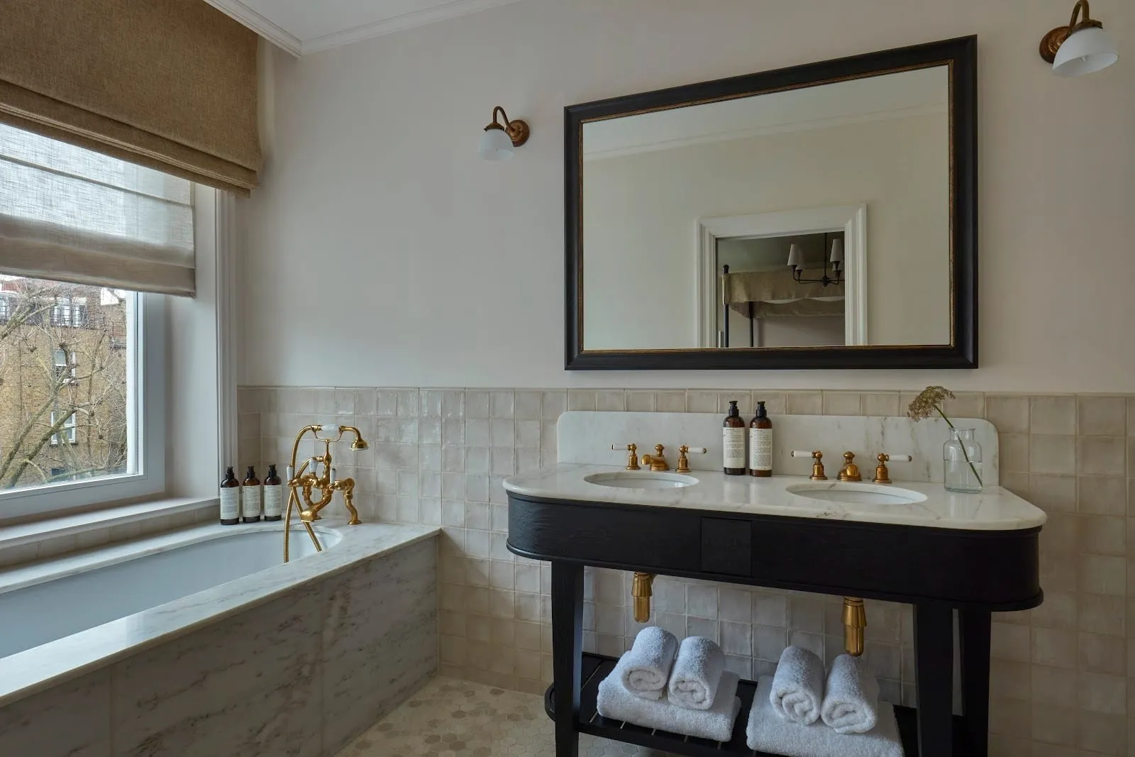 A Templeton Garden bathroom with a marble-panelled bath and brass taps under a window, and a black double-basin washstand with a marble top and rolled towels on the shelf below