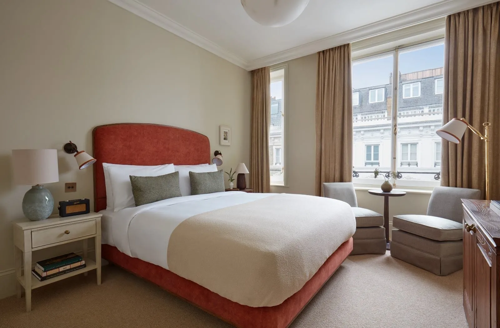 A Templeton Garden bedroom with a rust-red velvet headboard, a cream boucle throw, a small radio on the bedside table and two armchairs by a tall window looking across at white stucco houses