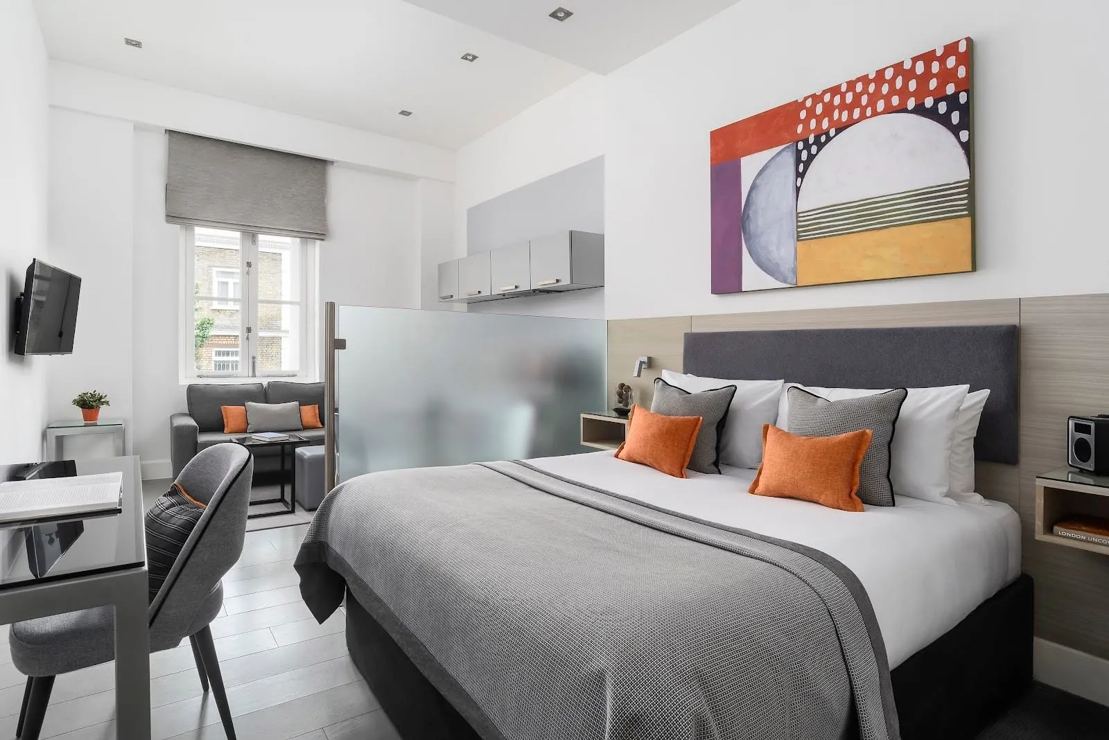 A Templeton Place studio with a double bed under an abstract painting, a frosted glass screen hiding the kitchenette and its grey wall cupboards, a sofa by the window and a desk