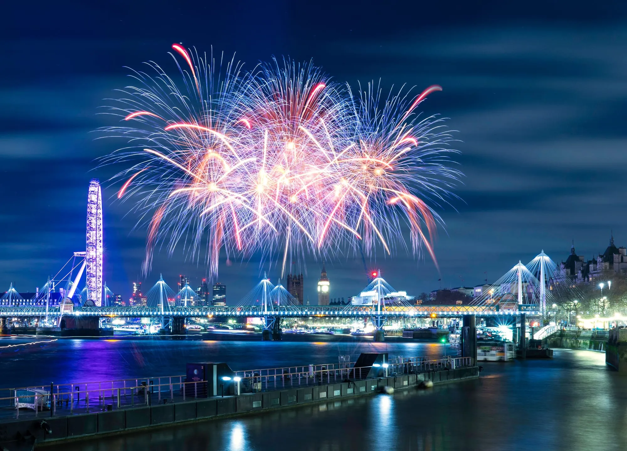 Fireworks bursting in red and blue over the Thames at night, with the London Eye, the Golden Jubilee Bridges and the Elizabeth Tower lit below