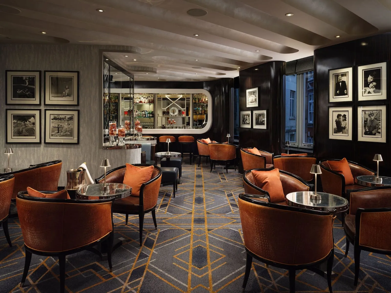 Tan leather armchairs with orange cushions around small round tables on a grey-and-gold geometric carpet, with framed black-and-white photographs on the walls and a curved back bar at the far end